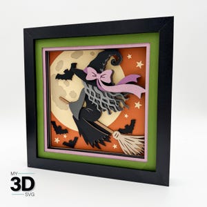 Peut inclure: Une œuvre d'art en papier superposé dans un cadre noir représentant une sorcière aux cheveux gris avec un nœud rose volant sur un balai devant une grande lune jaune, un ciel orange et des chauves-souris noires. Le texte MY 3D SVG est visible.