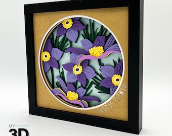 May include: A framed paper craft of purple and yellow flowers with green leaves. The flowers are arranged in a circle and the text "Pasque Flower" is written at the bottom of the frame.
