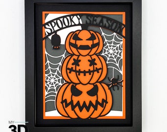 May include: A black picture frame with a white background and a black and orange paper cut-out design of three stacked jack-o'-lanterns with the text "Spooky Season" above them.