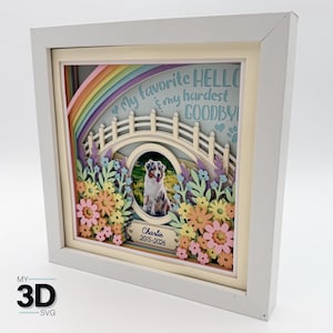 Op de afbeelding: Een ingelijst 3D-papierkunstwerk met een regenboog, brug en bloemenontwerp. Het kunstwerk bevat een foto van een hond genaamd Charlie, met de tekst "My favorite HELLO & my hardest GOODBYE." De lijst is wit.