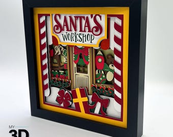 May include: A framed 3D papercraft scene of Santa's Workshop. The scene is decorated with red and white candy canes, a red and green wreath, and a variety of toys. The text "Santa's Workshop" is displayed in a yellow banner.