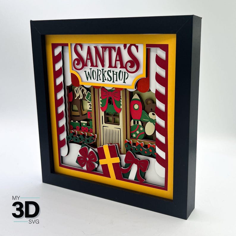 May include: A framed 3D papercraft scene of Santa's Workshop. The scene is decorated with red and white candy canes, a red and green wreath, and a variety of toys. The text "Santa's Workshop" is displayed in a yellow banner.