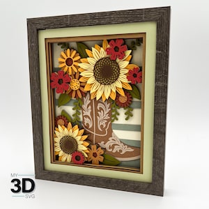 May include: A framed 3D layered artwork showing a brown cowboy boot overflowing with sunflowers and other flowers in yellow, red, and brown. The background has a striped pattern. The frame is dark grey.