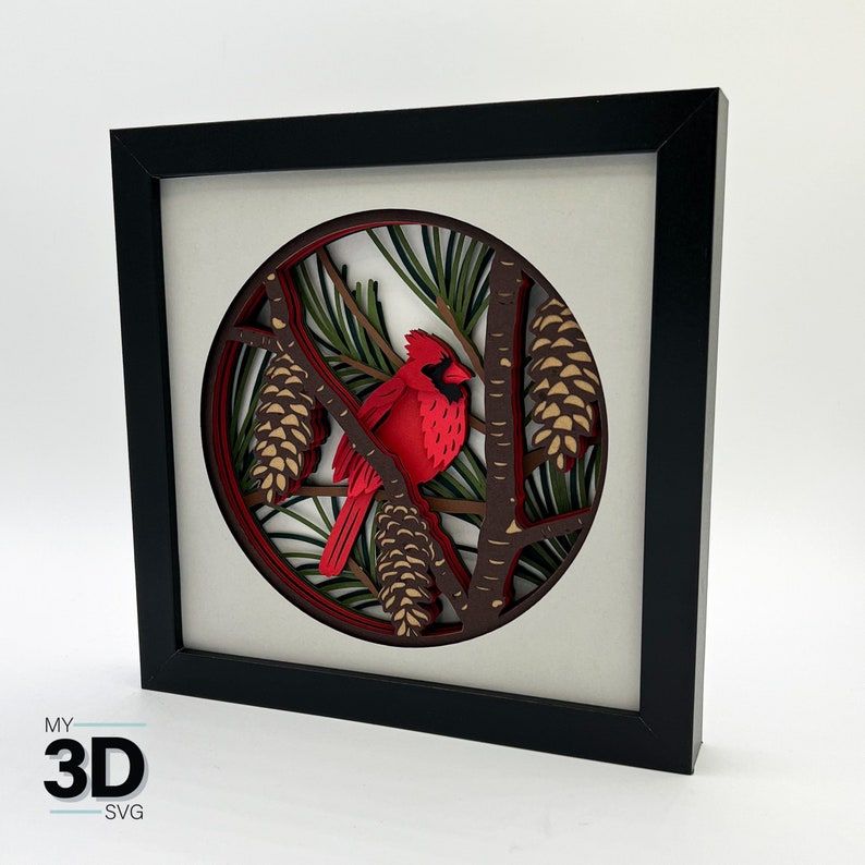 Puede incluir: Una obra de arte de papel recortado en 3D en un marco negro. La obra de arte presenta un cardenal rojo posado en una rama con pi&ntilde;as y hojas verdes. La obra de arte est&aacute; en capas y crea un efecto 3D.