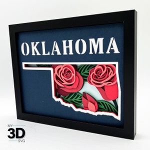 May include: A 3D paper craft of the state of Oklahoma with red roses and green leaves. The state outline is white and the background is blue. The word "OKLAHOMA" is cut out of the blue background.