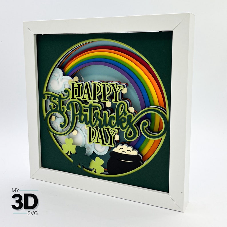 May include: A white frame with a green background and a layered paper craft design. The design features a rainbow, clouds, shamrocks, a pot of gold, and the text "Happy St. Patrick's Day".