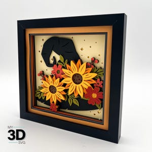 May include: A framed 3D paper art piece featuring a black witch's hat decorated with sunflowers and other flowers in shades of orange, yellow, and red. The artwork is set against a light beige background with star accents, within a black frame.