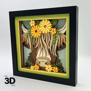 Puede incluir: Obra de arte en papel en capas enmarcada, que representa una vaca de las Highlands adornada con una corona floral. El marco es negro, con un passe-partout verde y amarillo. La vaca está representada en tonos marrones, crema y beige, con flores amarillas y hojas verdes.