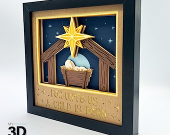 Puede incluir: Una artesanía de papel en 3D en capas de una escena de la natividad con un bebé Jesús en un pesebre, una estrella arriba y el texto "For unto us a child is born".