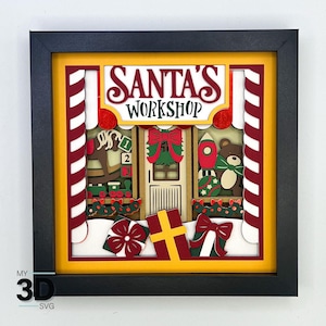 May include: A 3D layered paper craft of Santa's Workshop. The scene features a red and white striped candy cane, a rocking horse, a toy rocket, a teddy bear, and a door with a wreath. The sign above the door reads "Santa's Workshop".
