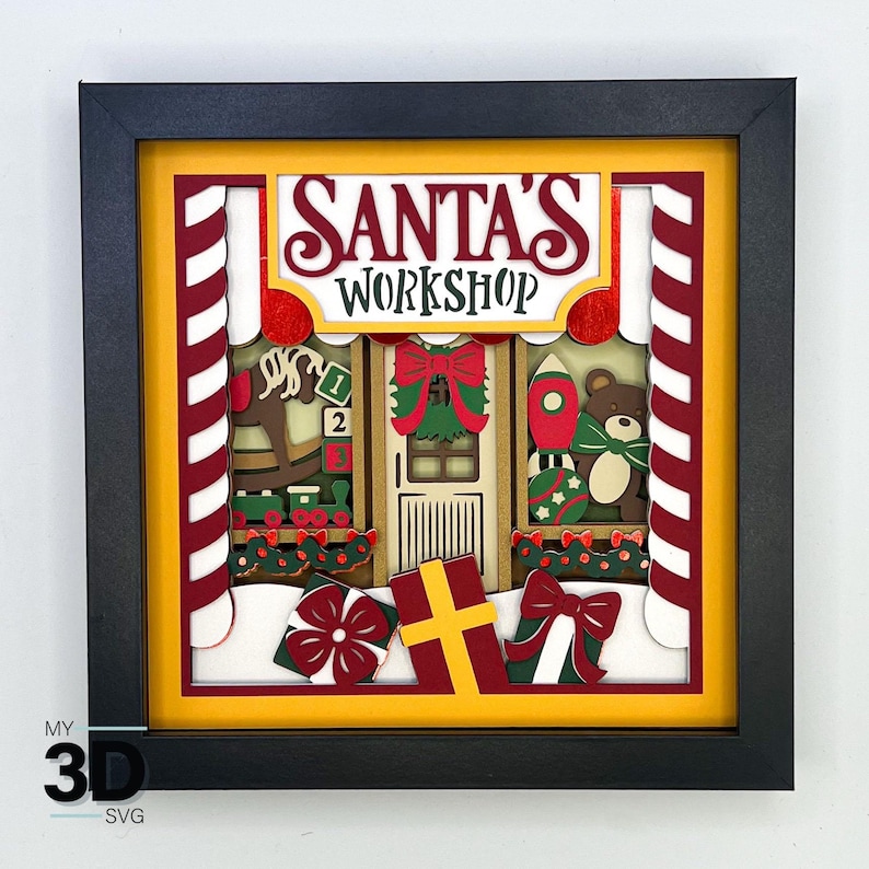 May include: A 3D layered paper craft of Santa's Workshop. The scene features a red and white striped candy cane, a rocking horse, a toy rocket, a teddy bear, and a door with a wreath. The sign above the door reads "Santa's Workshop".