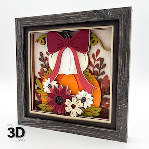 May include: A 3D layered art piece featuring a pumpkin design. The pumpkin is orange with a white top and a burgundy bow. It is adorned with sunflowers and framed in a dark gray wooden frame. The text "MY 3D SVG" is in the lower left corner.