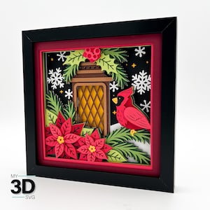 May include: A framed 3D paper art piece featuring a winter scene. The artwork includes a red cardinal, poinsettias, a lantern, snowflakes, and evergreen branches. The frame is black with a red matting, and the background is black.