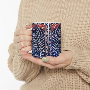 May include: A ceramic mug featuring a dark blue background with a repeating pattern of stylized trees with white and red flowers. The design includes a vertical border of small floral motifs and a red arched top section with white leaf details.