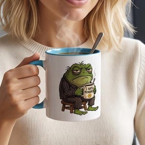 May include: A white ceramic mug with a light blue interior and handle, featuring a graphic of a grumpy green toad wearing dark striped pajamas, sitting on a wooden stool while holding a smaller mug labeled I HATE MCNOADS with a sad face icon.