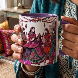 May include: A white ceramic mug with a purple handle and interior, featuring a colourful illustration of four figures in traditional South Asian attire dancing under festive bunting. The design includes vibrant pink, orange, and green patterns.