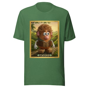 Op de afbeelding: Groen t-shirt met een ingelijste afbeelding van een aardappelvormig personage met bruin haar, een vest en het woord "TATERS" onderaan. Het personage staat tegen een landschap met een pad en glooiende heuvels.