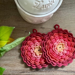 Könnte beinhalten: Zwei gehäkelte Topflappen in Form einer Blume. Die Topflappen sind aus einem Farbverlaufsgarn in Rot, Rosa und Orange gefertigt.