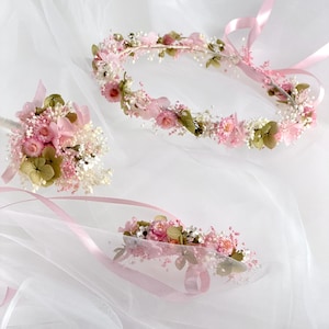 May include: A bridal set featuring a floral crown, a small bouquet, and a wrist corsage. The set is composed of pink and white flowers with green accents, and pink ribbons. The items are arranged on a white, sheer fabric.