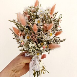 May include: A small bouquet of dried flowers in shades of white and pink, tied with a white lace ribbon. The bouquet includes daisies, baby's breath, and dried grasses. The text "Baby bouquet Alice" is written below the bouquet.