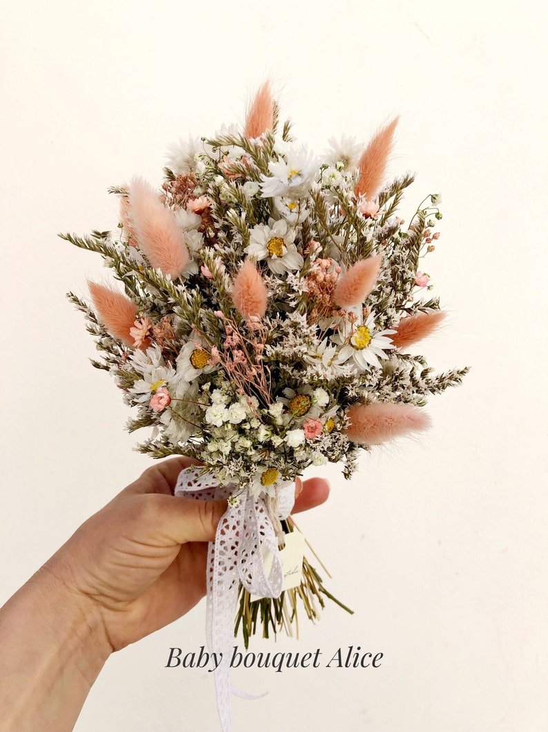 May include: A small bouquet of dried flowers in shades of white and pink, tied with a white lace ribbon. The bouquet includes daisies, baby's breath, and dried grasses. The text "Baby bouquet Alice" is written below the bouquet.