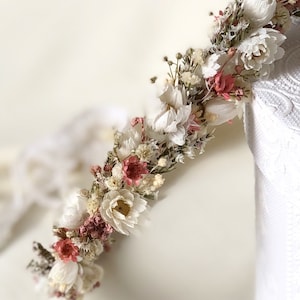 May include: A white and blush dried flower crown with a delicate, rustic aesthetic. The crown is made with a variety of dried flowers, including white baby's breath, blush pink flowers, and green foliage.