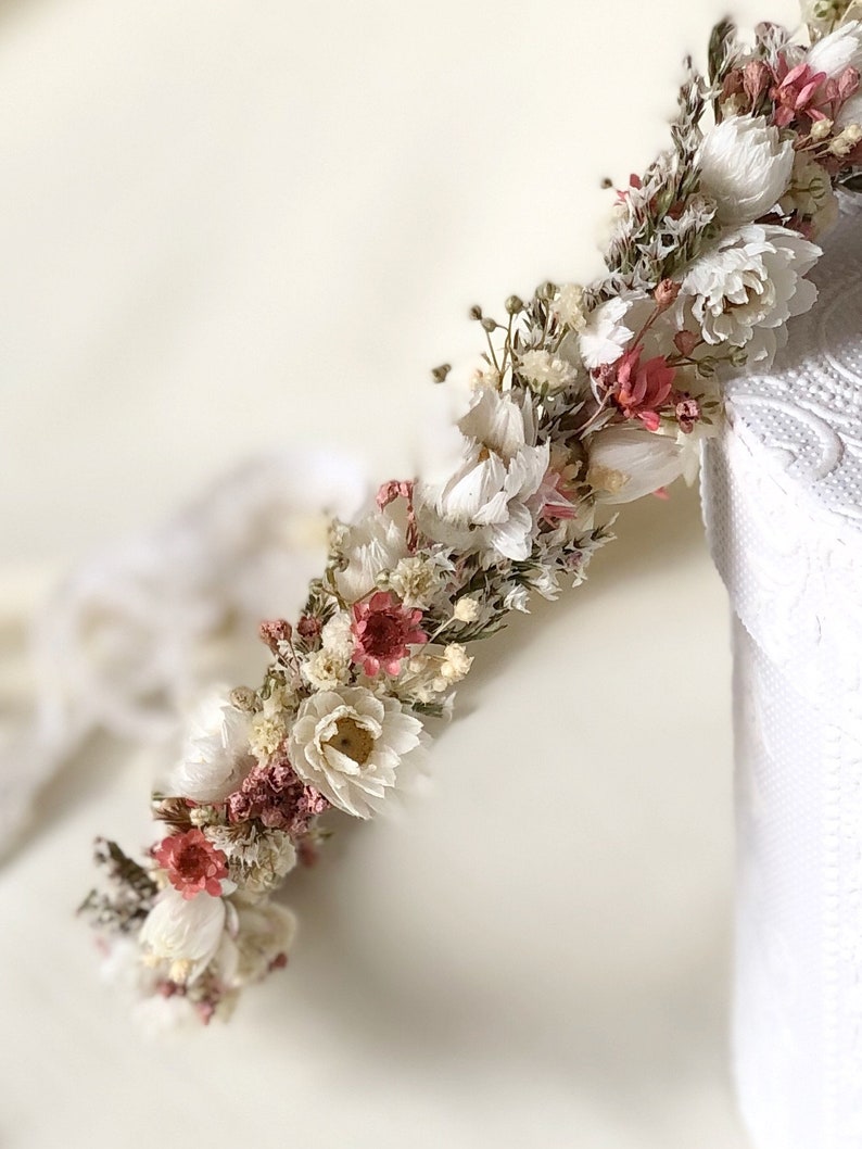 May include: A white and blush dried flower crown with a delicate, rustic aesthetic. The crown is made with a variety of dried flowers, including white baby's breath, blush pink flowers, and green foliage.