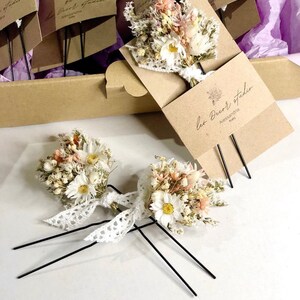 May include: Two hair clips featuring dried flower bouquets, with white daisies, pink flowers, and greenery. The bouquets are tied with white lace and attached to black metal hair pins. The text "Alice" is printed on the white surface below the hair clips.