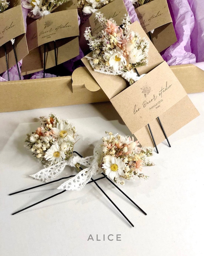 May include: Two hair clips featuring dried flower bouquets, with white daisies, pink flowers, and greenery. The bouquets are tied with white lace and attached to black metal hair pins. The text "Alice" is printed on the white surface below the hair clips.