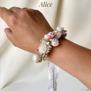 May include: A white and pink floral bracelet with a white lace ribbon. The bracelet is made with dried flowers and is tied around a wrist.