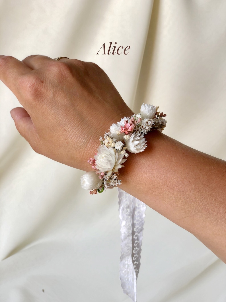 May include: A white and pink floral bracelet with a white lace ribbon. The bracelet is made with dried flowers and is tied around a wrist.