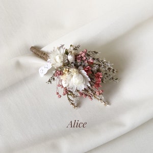 May include: A small bouquet of dried flowers in shades of white and pink, tied with twine and a small piece of lace. The bouquet is arranged on a white fabric background. The text "Alice" is written below the bouquet.