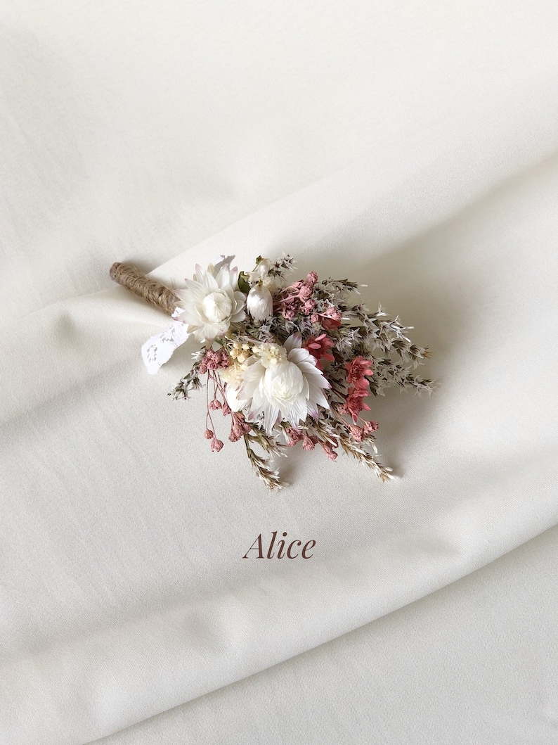 May include: A small bouquet of dried flowers in shades of white and pink, tied with twine and a small piece of lace. The bouquet is arranged on a white fabric background. The text "Alice" is written below the bouquet.