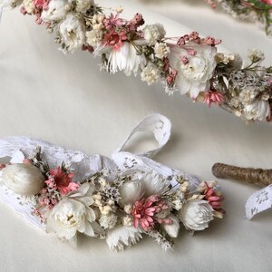 May include: A delicate flower crown and matching wristlet made with dried flowers in shades of white and pink. The crown is made with a variety of flowers, including small white flowers, pink flowers, and green foliage. The wristlet is made with a similar variety of flowers and is attached to a white lace band.