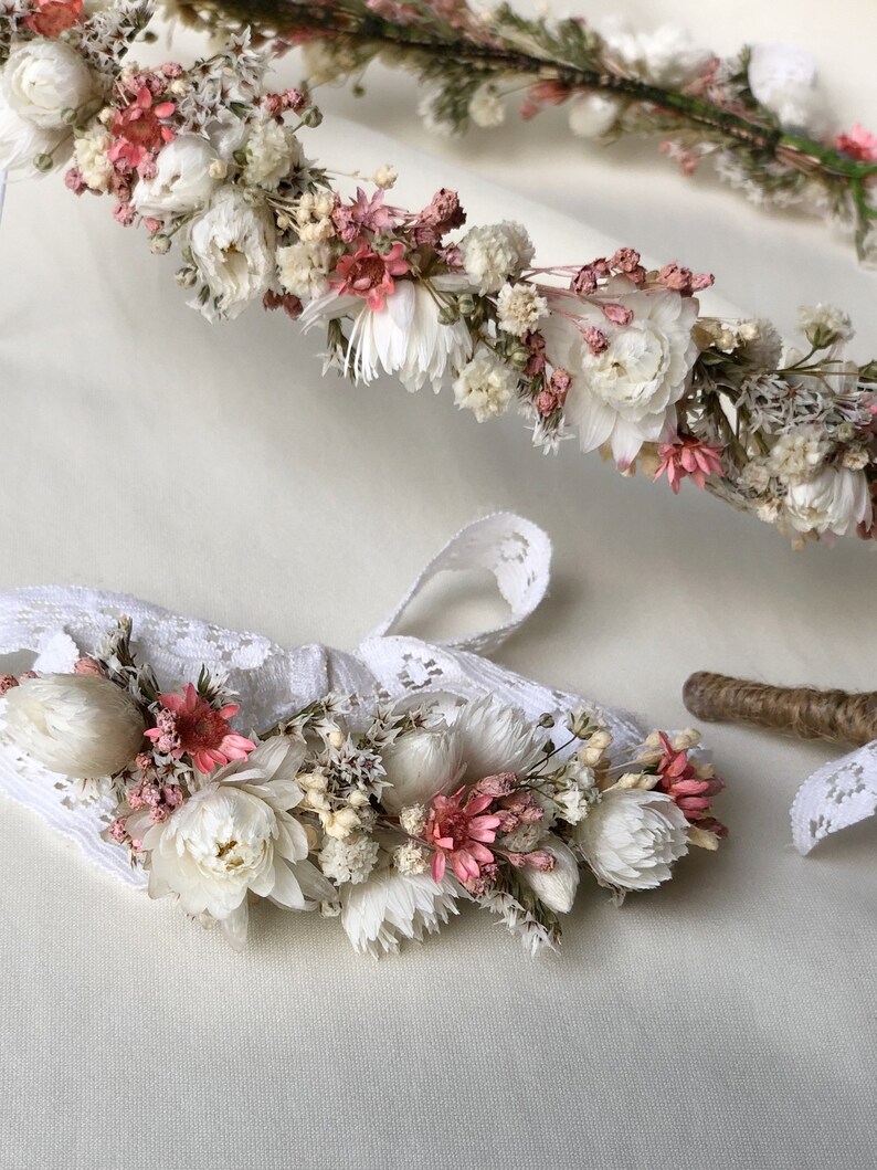 May include: A delicate flower crown and matching wristlet made with dried flowers in shades of white and pink. The crown is made with a variety of flowers, including small white flowers, pink flowers, and green foliage. The wristlet is made with a similar variety of flowers and is attached to a white lace band.