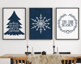 Puede incluir: Tres impresiones enmarcadas con un esquema de color azul y blanco. La primera impresión presenta una silueta blanca de un árbol de Navidad con el texto "Wishing you a very merry Christmas". La segunda impresión presenta un copo de nieve blanco sobre un fondo azul oscuro. La tercera impresión presenta una corona blanca con el texto "Tis the season" y una campana.