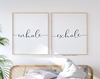 Puede incluir: Dos impresiones enmarcadas con las palabras "inhale" y "exhale" en una elegante escritura. Las impresiones se muestran sobre una cama con un edredón azul claro y una cabecera tejida. Un banco y plantas en macetas completan la serena escena del dormitorio.