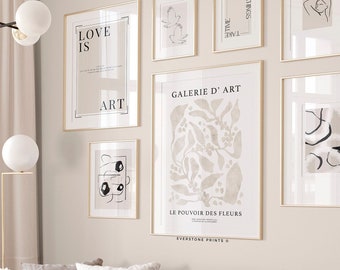 Puede incluir: Un interior de sala de estar con un sofá beige y almohadas blancas. Un ramo de peonías blancas se encuentra sobre una mesa de café blanca. La pared está decorada con una pared de galería de impresiones de arte en blanco y negro con marcos dorados. Las impresiones presentan diseños abstractos, texto y motivos florales. El texto en las impresiones dice "Love is Art", "Galerie d'Art", "Le Pouvoir des Fleurs", "Good Time", "Goodness", y "Everstone Prints".