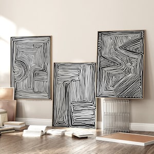 Puede incluir: Tres impresiones de arte abstracto en blanco y negro con marcos dorados colgadas en una pared blanca. Las impresiones están dispuestas en línea vertical, con la más grande en la parte superior y la más pequeña en la parte inferior. Las impresiones son todas de diseños diferentes, pero todas tienen un estilo similar. Las impresiones están colgadas en una pared blanca con un suelo de madera.