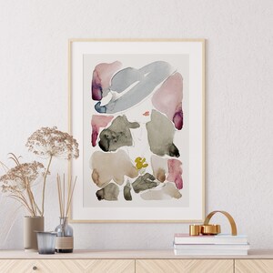 May include: A framed abstract watercolour painting with pink, grey, and brown shapes. The painting is on a white wall above a wooden shelf with a vase of dried flowers, a glass vase, and a gold metal decorative object.