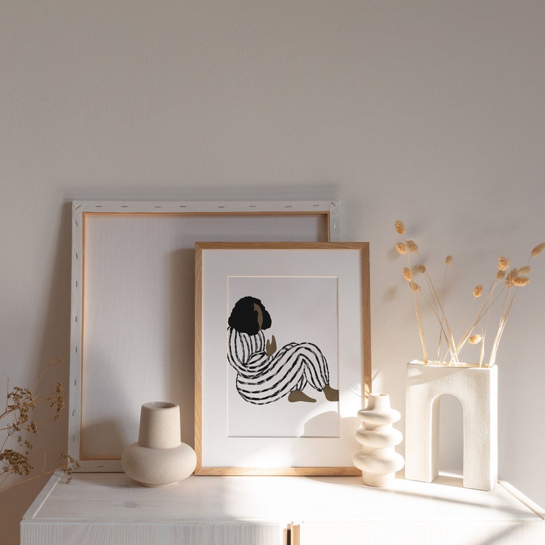 May include: A framed illustration of a person in a black and white striped outfit sitting with their legs crossed. The illustration is in a light wood frame and is sitting on a white shelf with a white vase and a white ceramic object.