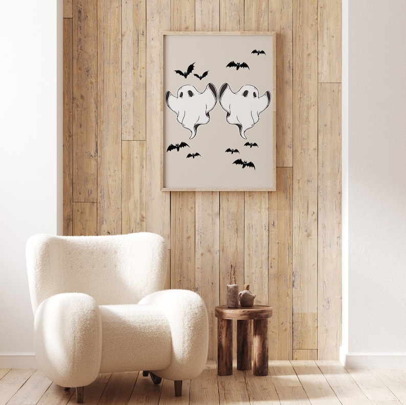 May include: A framed print with a beige background featuring two white ghosts with black eyes and outstretched arms. The ghosts are surrounded by black bats. The print is hanging on a wood wall.