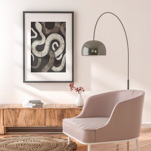 May include: A framed print of a black and white abstract snake design hangs on a white wall above a wooden console table. A pink armchair sits in front of the table with a small vase of red berries on the table.