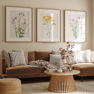 May include: Three framed botanical prints of different flowers in a living room setting. The prints are hung on a light beige wall above a brown leather sofa with throw pillows and a blanket. A wooden coffee table sits in front of the sofa.