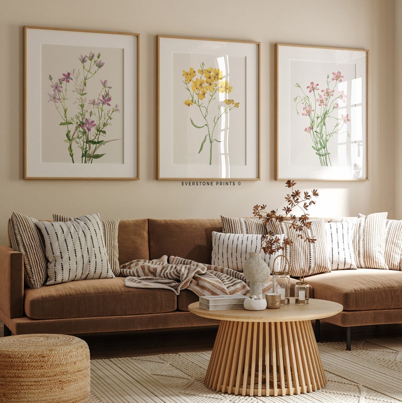 May include: Three framed botanical prints of different flowers in a living room setting. The prints are hung on a light beige wall above a brown leather sofa with throw pillows and a blanket. A wooden coffee table sits in front of the sofa.