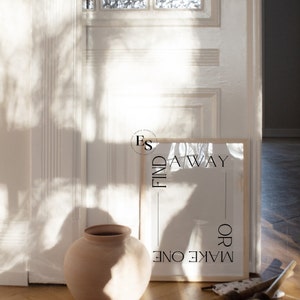 May include: A framed print with the text "FIND A WAY OR MAKE ONE" in a white frame on a wooden floor. The print is leaning against a white door with a shadow cast on the wall.