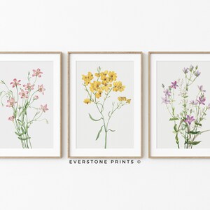 May include: Three framed botanical prints featuring watercolor illustrations of pink, yellow, and purple flowers. The prints are displayed on a white wall with a light wood frame.