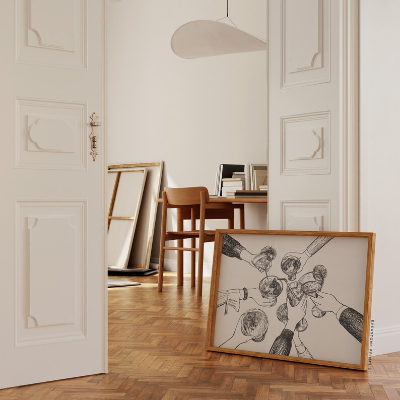 Op de afbeelding: Een ingelijste zwart-witte illustratie van handen die glazen met drankjes omhoog houden. De illustratie is op een lichtbeige achtergrond. De lijst is van hout en heeft een lichtbruine afwerking. De tekst "EVERSTONE PRINTS" is gedrukt in de rechterbenedenhoek van de lijst.