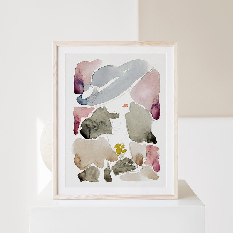 May include: An abstract watercolour painting in a light wood frame. The painting features a woman's face with a large hat, surrounded by abstract shapes in shades of pink, purple, grey, and brown.