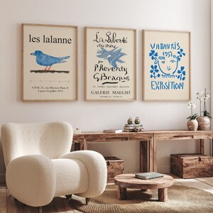 May include: Three framed art prints with a white background and blue text and illustrations. The left print features a blue bird with the text "les lalanne" and "CNAC 11, rue Berryer Paris B 5 juin-13 juillet 1975". The center print features a blue illustration of a bird with the text "La Libert&eacute; des mers par P.Reverdy G.Braqus. ET LIVRES ILLUSTRES RECENTS GALERIE MAEGHT". The right print features a blue illustration of a face with flowers around it and the text "VALAVRIS 1957 EXPOSITION".